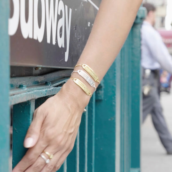 A girl grazes her hand against a green subway entrance, wearing 5 of our engraved bracelets in gold and silver.